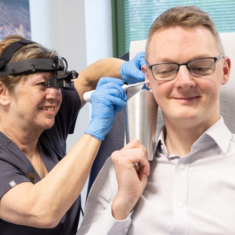 Female Audiologist wearing specialist head loupes whilst conducting ear irrigation on a male patient. Male patient is sitting upright.