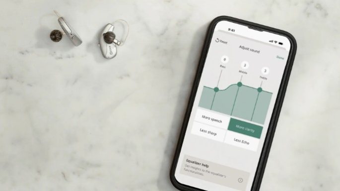A pair of silver Widex Allure hearing aids sitting on a counter top, next to a smartphone showing the Widex App.