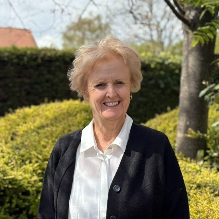 All About Hearing Customer Care Team member, Dawn Mucha, smiling outside with green foliage behind her.