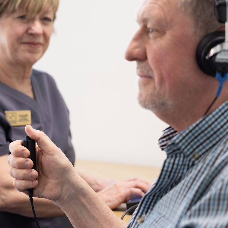 Male patient holding a black response button whilst carrying out a hearing assessment. Female Audiologist in background.